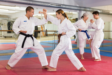 Woman and man in white kimono and belts sparring during karate training. Elderly woman and young man sparring in background. - obrazy, fototapety, plakaty
