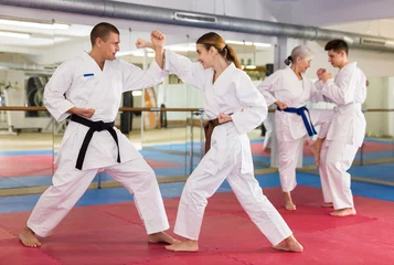 Fotobehang Woman and man in white kimono and belts sparring during karate training. Elderly woman and young man sparring in background. © JackF