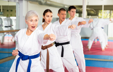 Concentrated aged woman in kimono practicing punches in gym during group martial arts workout....