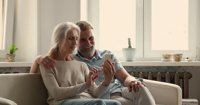 Happy mature carefree couple sit on sofa in living room at home have fun use modern smartphone. New cool mobile app usage, remote easy comfort services for older citizen, free time on internet concept
