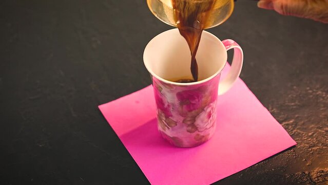 espresso being poured into floral cup and then steamed, frothed milk over top to make a latte, cappuccino.  Pink square napkin under cup on a black counter top. 