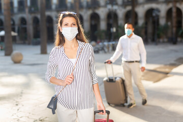 Portrait of young female traveler in medical mask strolling with luggage along city street on spring day. New lifestyle concept during coronavirus..