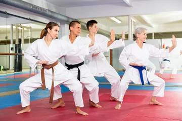 Fotobehang Group of focused people wearing white kimonos standing in attacking stance, practicing movements of close combat punches in training room during martial arts workout.. © JackF