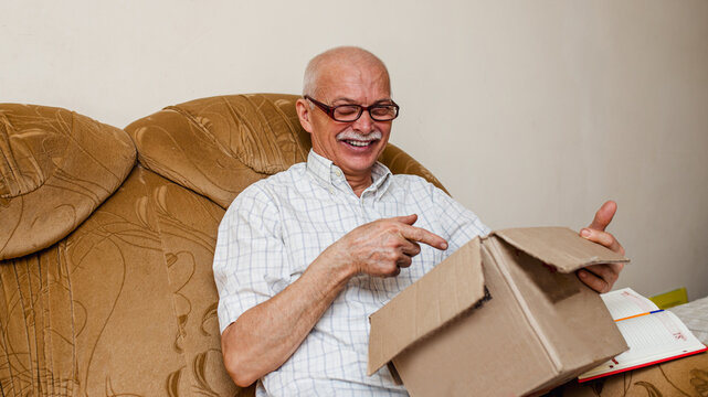 Happy mature elderly man inspects received parcel. old man sits on vintage sofa in living room. Online order for fast courier delivery for senior citizen. Family support, care. Quarantine, isolation.