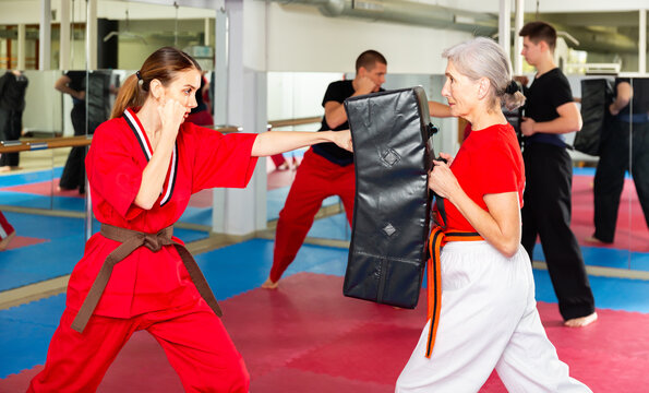 Concentrated young woman in red kimono launching blows on punch shield during martial arts training in gym with aged female partner