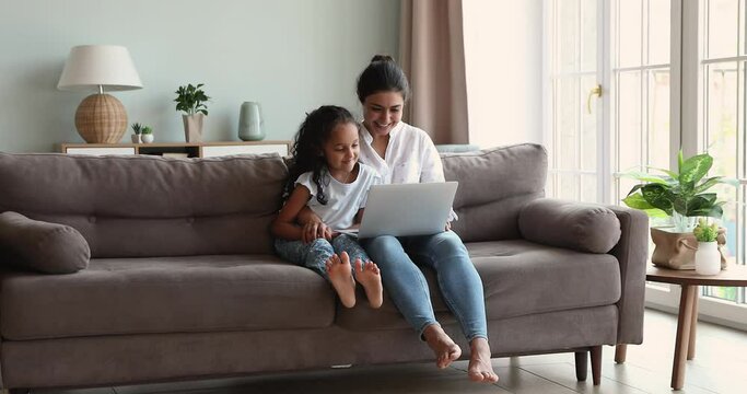 Indian woman her little 6s daughter spend time at home enjoy internet fun, sit on couch with laptop, watch educational vlog on-line content. Children gen Z and development using modern tech concept