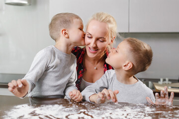 A young mother and two sons cook in a large and bright kitchen. Mom teaches children to cook cookies. Sons kiss your mom. The photo shows the value of family, love and care.