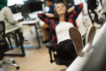 Casual business woman taking a break with legs on her table while working on desktop computer in modern open plan startup office interior. Selective focus 