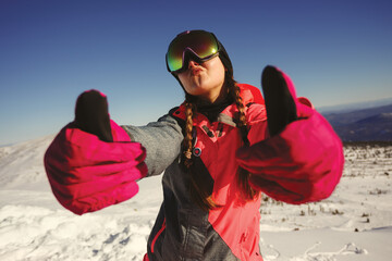 Young beautiful girl snowboarder with mask on her face holds thumbs up.