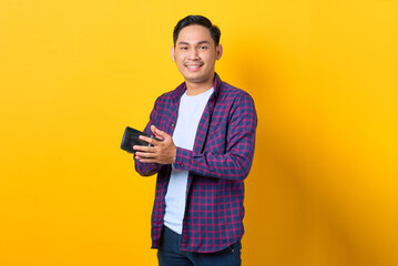 Smiling young Asian man in plaid shirt holding wallet isolated on yellow background