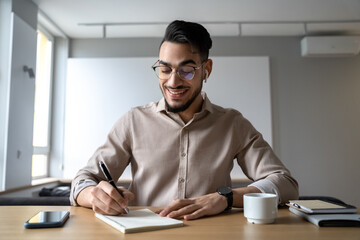 Happy smiling man sitting at desk writing in notepad listen online course