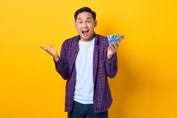 Surprised young Asian man in plaid shirt holding money banknotes isolated on yellow background