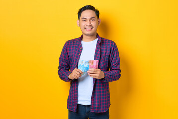 Smiling happy young Asian man in plaid shirt holding money banknotes isolated on yellow background