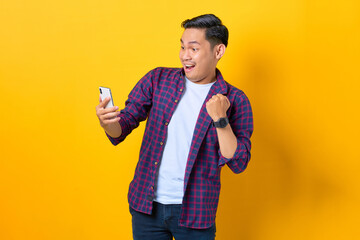 Excited young Asian man in plaid shirt using smartphone and doing winner gesture isolated on yellow...