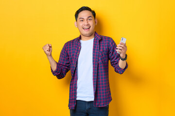 Excited young Asian man in plaid shirt using smartphone and doing winner gesture isolated on yellow background