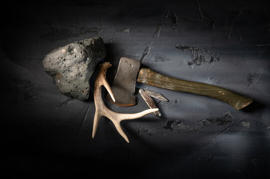 Rugged stone rock with vintage camping hatchet axe and deer antler on textured dark gray surface.