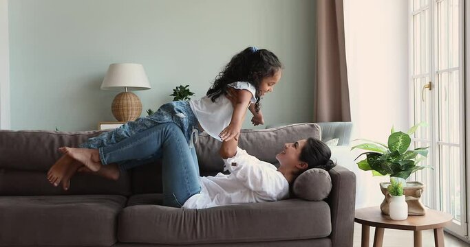 Young Indian mother lying on cozy sofa in living room raises her daughter in her arms play airplane, dream about traveling feel happy, enjoy active games, playtime at modern home. Fun, leisure concept