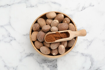 Scoop with nutmeg powder and seeds in bowl on white marble table, top view