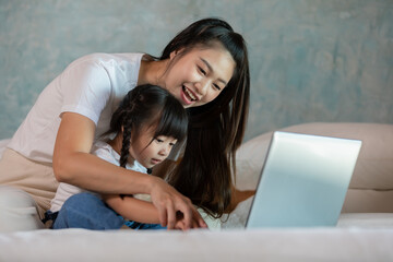 Fototapeta premium Beautiful Asian mom teaching her daughter to learn and using computer laptop for preschool or watching cartoons online at home.Happy mother relax with little girl having fun with laptop at home