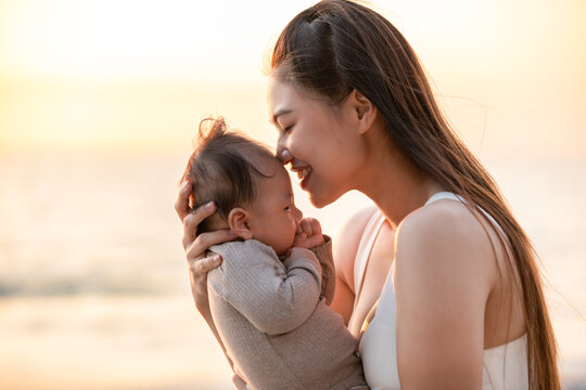 Beautiful Attractive Asian mom holding her baby newborn in hand and kissing on baby head sweet and lovely outdoor on the beach.Happy mother and infant baby looking together smile with natural sunset