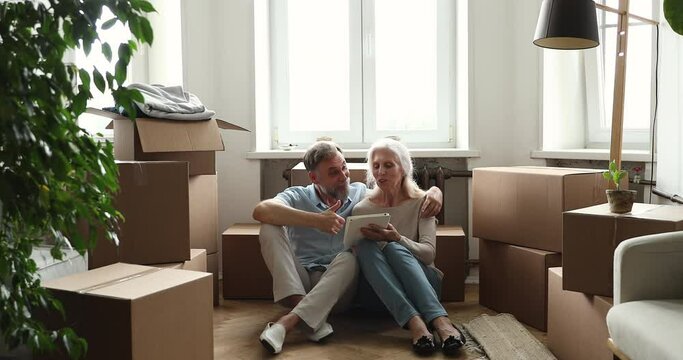 Older couple use digital tablet search repairs ideas on internet, buy furniture, interior items for flat sit on floor near heap of boxes with belongings. Retail services application, new house concept