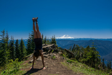 Adventurous athletic woman doing a handstand on a mountain top with Mount Rainier in the background.