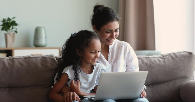 Indian mom and daughter sit on sofa use laptop watch cartoon, family movie on digital movie streaming platforms online services, discuss purchase on internet webstore. Ecommerce, leisure, tech concept