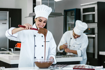latin woman pastry chef wearing uniform holding a bowl preparing delicious sweets chocolates at...
