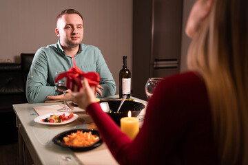 Happy young couple celebrating anniversary or valentine's day having romantic dinner at home table. A loving woman gives a gift box to her beloved man