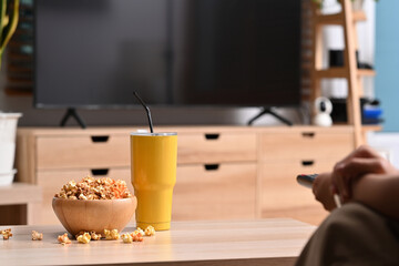 Young woman sitting on sofa with remote control while watching TV and eating popcorn.