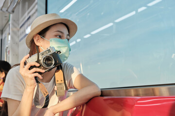 Beautiful Asian female tourist with face mask sits in a red seat, traveling by train, taking snapshot photo, transporting in suburb view, enjoy passenger lifestyle by railway, happy journey vacation.