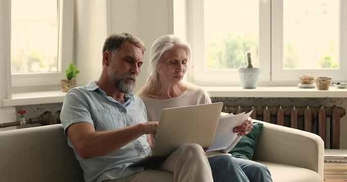 Mature spouses sit on couch at home with laptop learn received legal documents, sorting out papers, making online payment, discuss expenses, reviewing pension status, family budget management concept