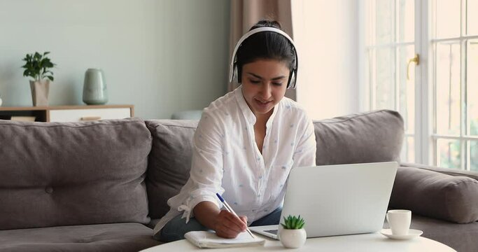 Indian student wear headphones sit at table studying writes notes in copybook, makes assignment, e-learning foreign language talk to online teacher. Virtual class, webinar, gain new knowledge concept