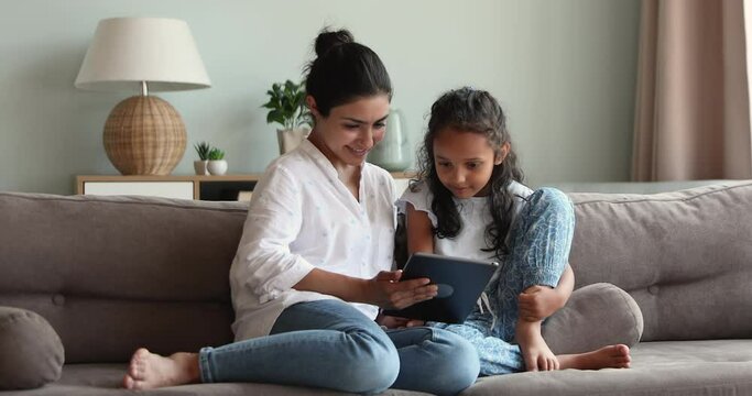 Loving Indian mother showing to little 6s daughter new cool application on digital tablet for development, family spend time at home use device, enjoy on-line fun. Generation z and modern tech concept