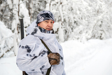 Portrait of a male hunter in a winter forest with a gun in a camouflage white suit. The concept of winter hunting.