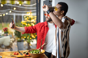 Two brightly dressed stylish guys taking selfie on phone, while having fun together at home. Concept of multiracial diverse gay couple and style. Caucasian and hispanic man having close relationship