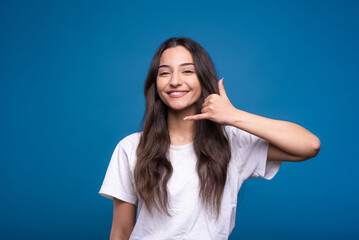Attractive caucasian or arab brunette girl in a white t-shirt showing a call gesture on a mobile phone isolated in on a blue studio background.