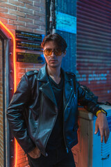 Portrait of young stylish brunet caucasian man in black leather jacket and yellow sunglasses. Standing on the street near the wall in evening. 