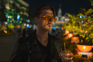 Portrait of young handsome brunet caucasian man in black leather jacket and yellow sunglasses. Standing on the street at night and holding ice coffee.