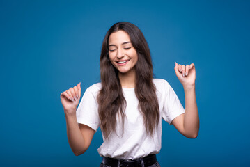 Attractive caucasian or arab brunette girl in a white t-shirt is dancing with her eyes closed and showing a winner gesture isolated on a blue studio background.