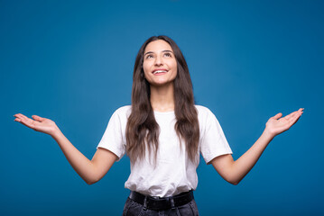 Young and attractive caucasian or arab brunette girl in white t-shirt giving thank to god and looking up isolated on blue studio background.