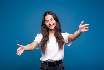 Young and attractive caucasian or arab brunette girl in a white t-shirt is positive and showing a hug and love gesture isolated on a blue studio background.