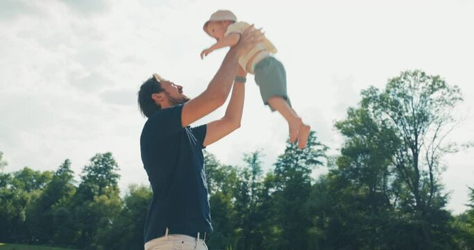 Playing with a child outdoors, by the water, lake, a man tosses a boy up and catches him, a father spends time with his son who is wearing a hat on head to protect him from the sun