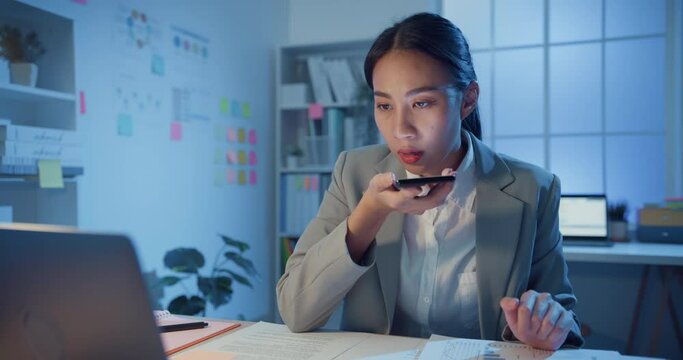 Young Asia cheerful professional woman sitting on desk with laptop record audio voice message on smartphone confirm detail in office at night. Lady business suit, Technology startup business concept.