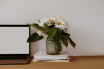Aesthetic home office desk workspace. Blank screen laptop computer with copy space. Daisy flowers bouquet in vase on table. Influencer lifestyle blog