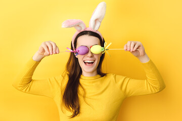Funny young caucasian woman covers eyes with two Easter eggs, wears fluffy bunny ears, dressed in sweater, model on yellow background with free space for promotion. Spring holiday and Easter concept