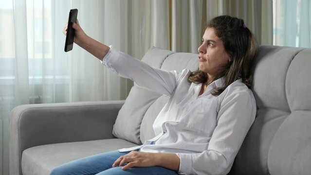 Cheerful disabled woman talks on videocall with friend via smartphone waving hand to greet companion sitting on comfortable sofa in room at home.