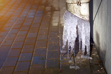 Drain pipe with dripping ice on sunny spring day, wet sidewalk. Ice in drainpipe, ice dam. Roof gutter with icicle. Downpipe on residential building with melting icicles in spring.