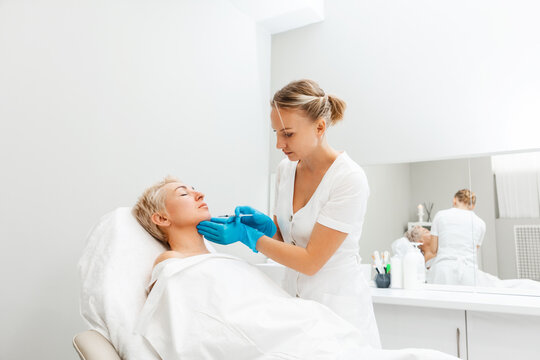 Professional cosmetologist in medical gloves with syringe injects filler in cheen. Adult woman getting injection in the cosmetology salon