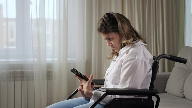 Young disabled woman with infantile cerebral paralysis tries to read news in mobile phone sitting in wheelchair near window at home side view.